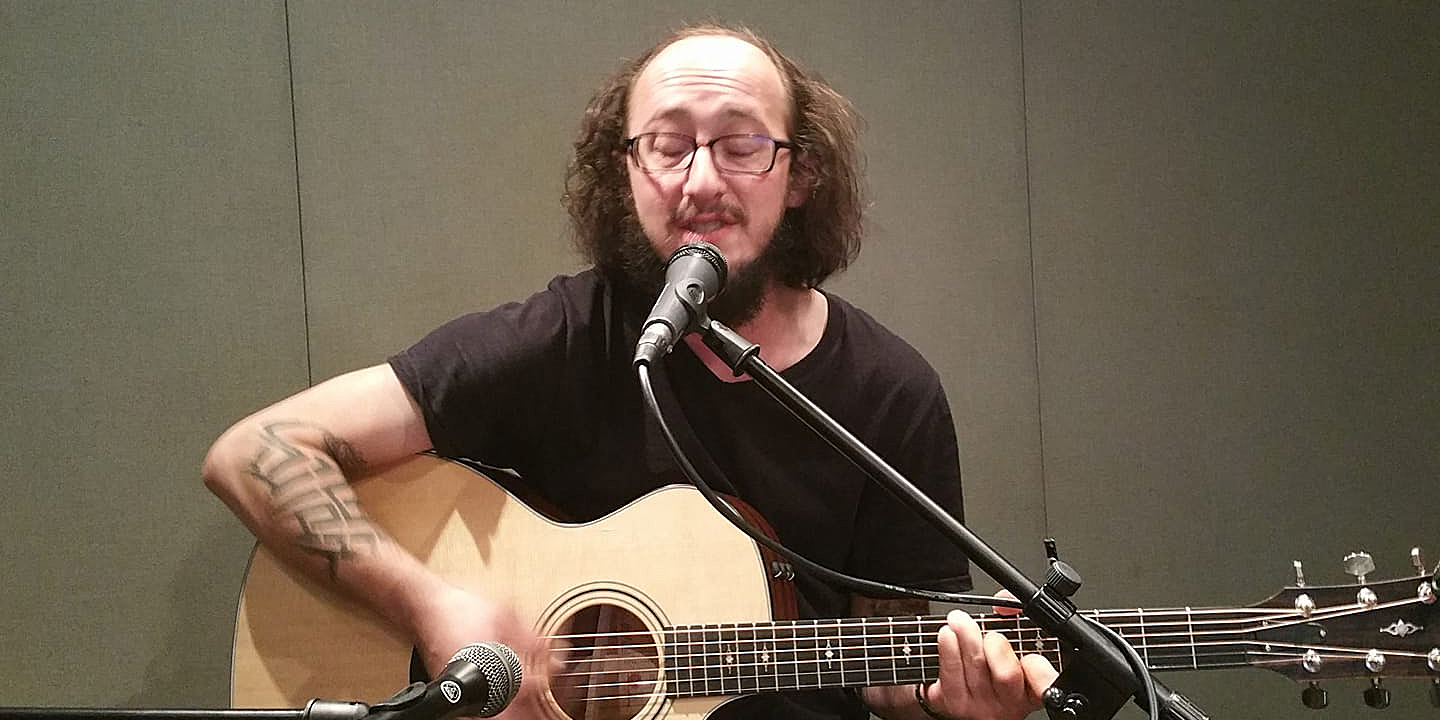 An adult playing acoustic guitar and singing into a microphone in a radio studio