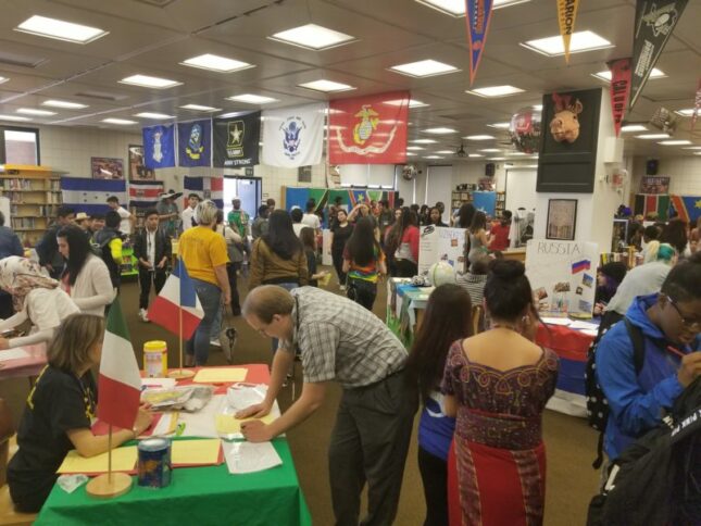 A large crowd of adults milling around a room with flags honoring the United States military and tables with student projects describing multiple foreign countries
