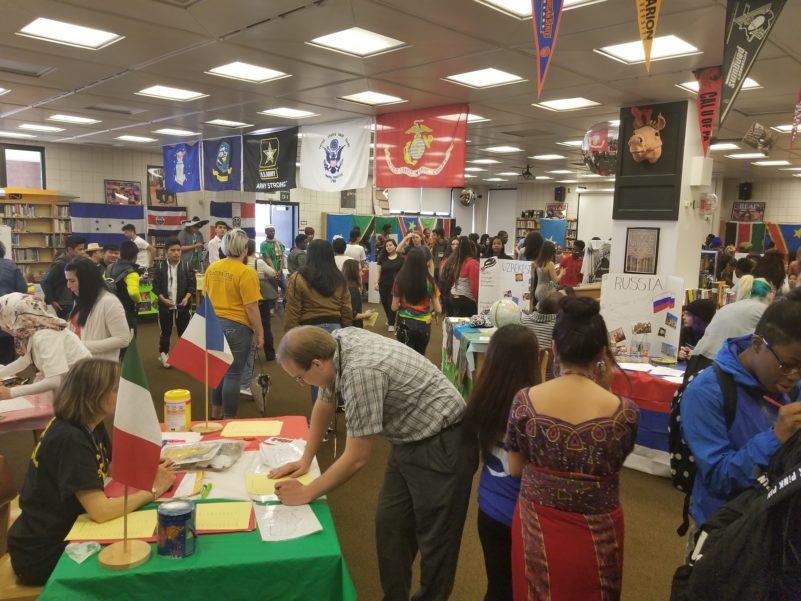A large crowd of adults milling around a room with flags honoring the United States military and tables with student projects describing multiple foreign countries
