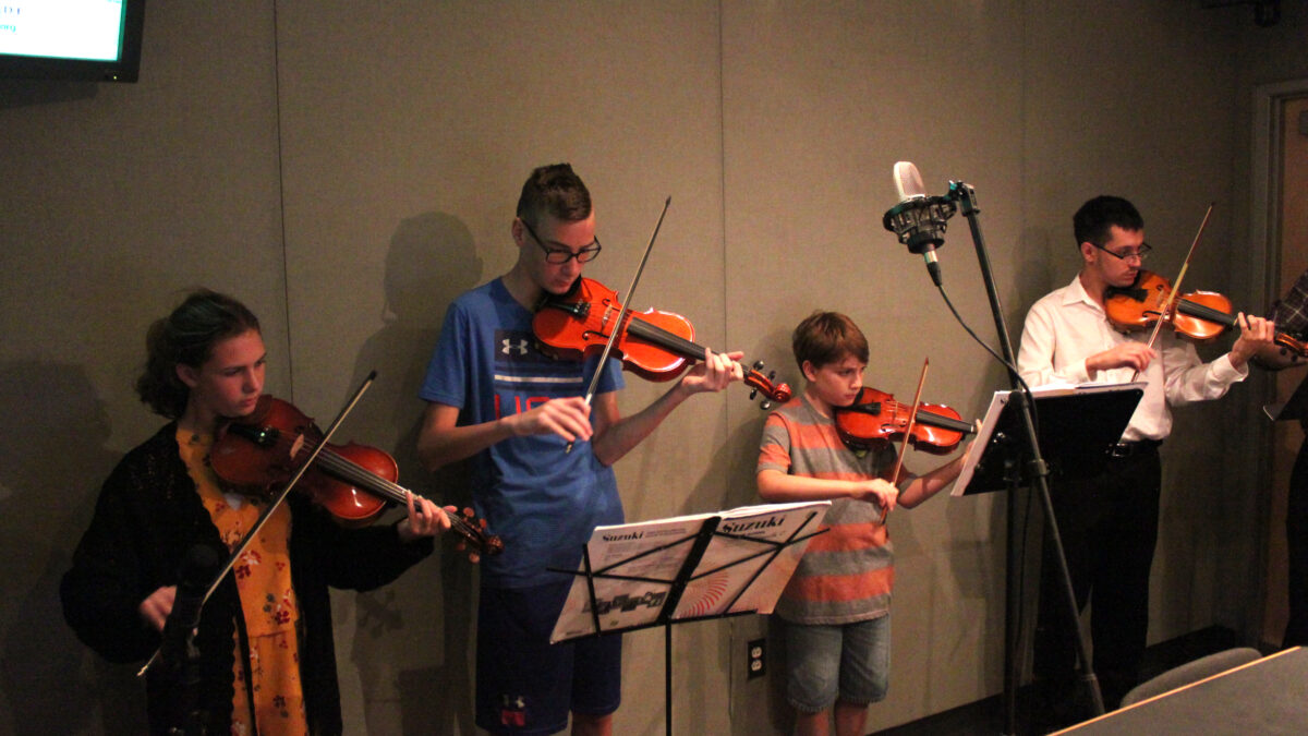 Four youth playing violins and looking at sheet music on music stands in a radio studio
