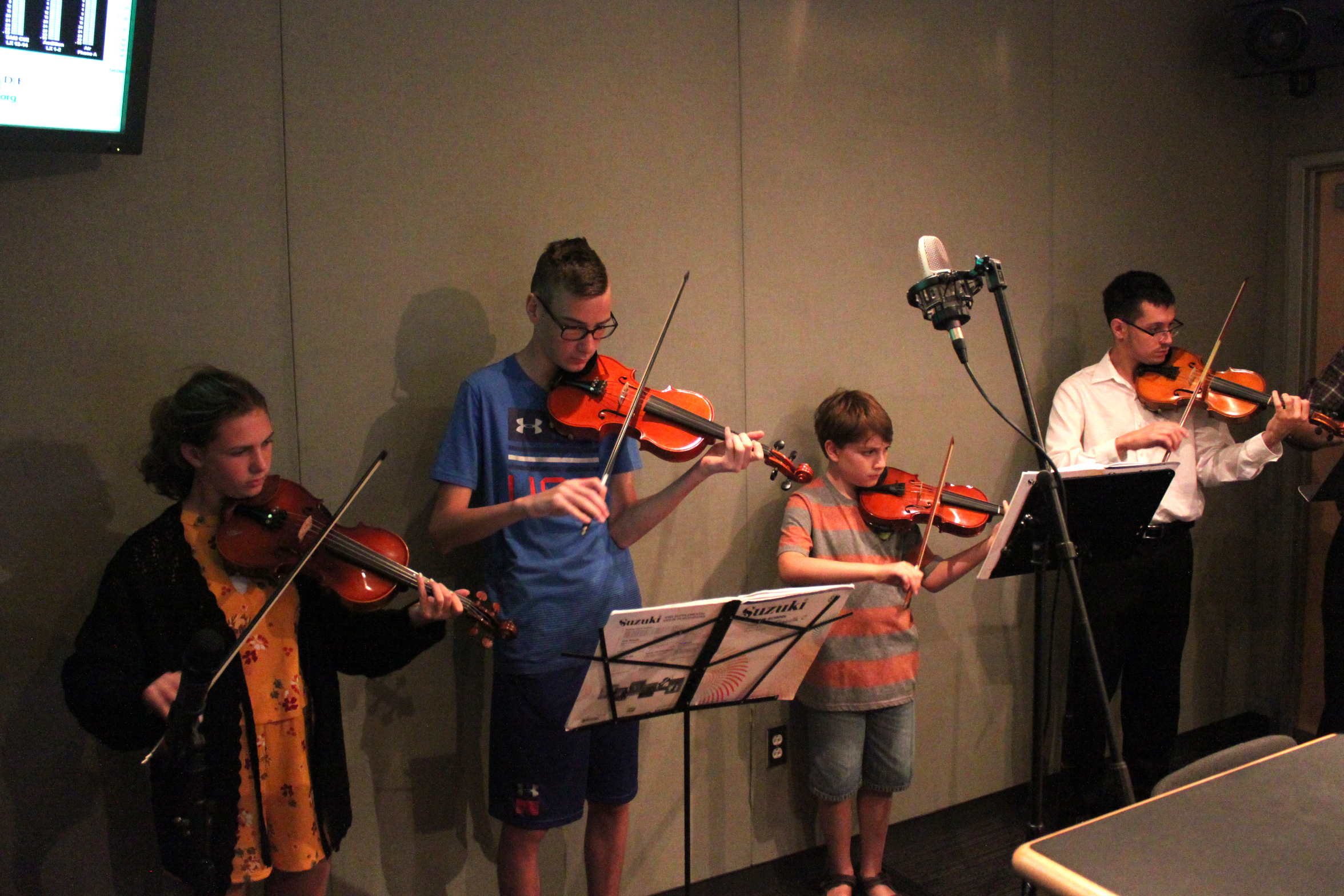 Four youth playing violins and looking at sheet music on music stands in a radio studio