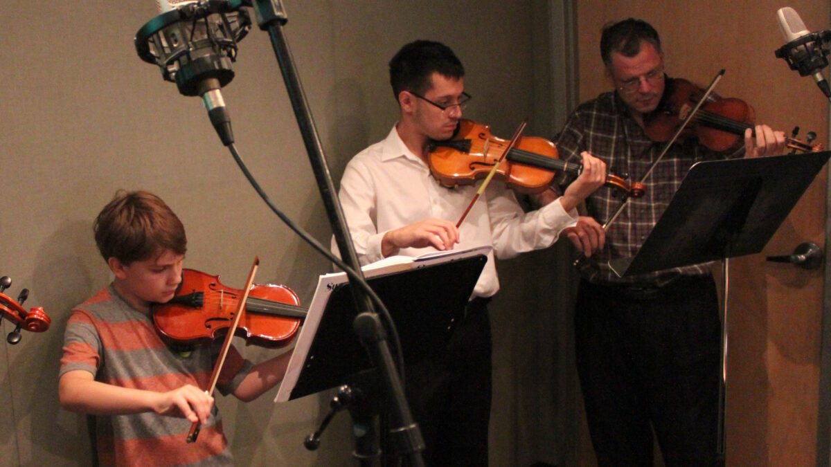 Two youth and one adult playing violins and looking at sheet music on music stands in a radio studio