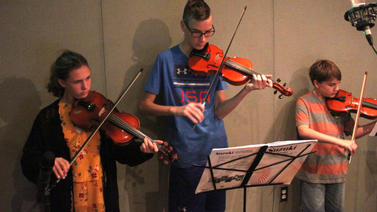 Three youth playing violins looking at sheet music on a music stand in a radio studio