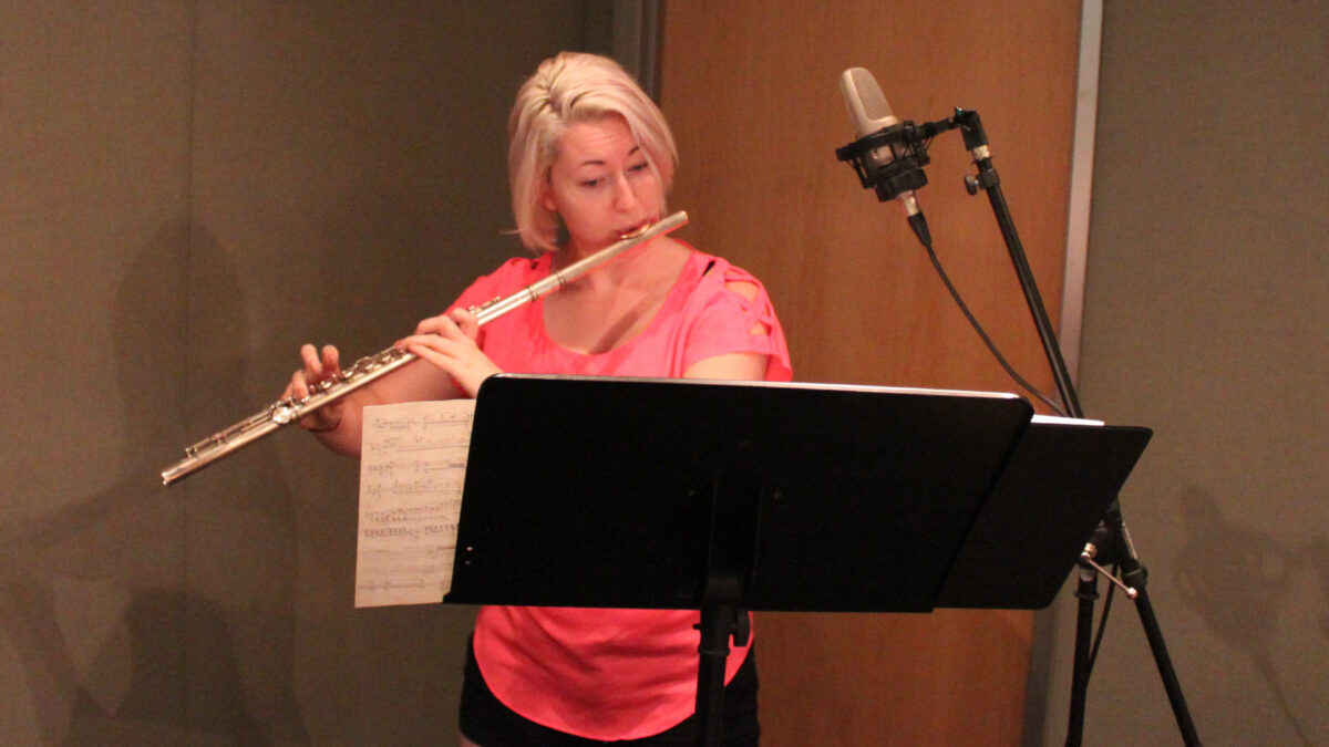 An adult playing flute looking at sheet music on a music stand in a radio studio