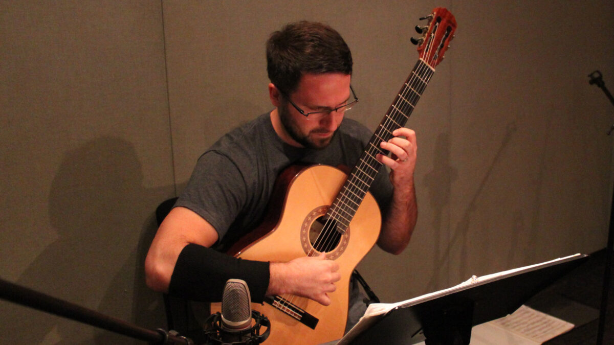 An adult playing acoustic guitar looking at sheet music on a music stand in a radio studio