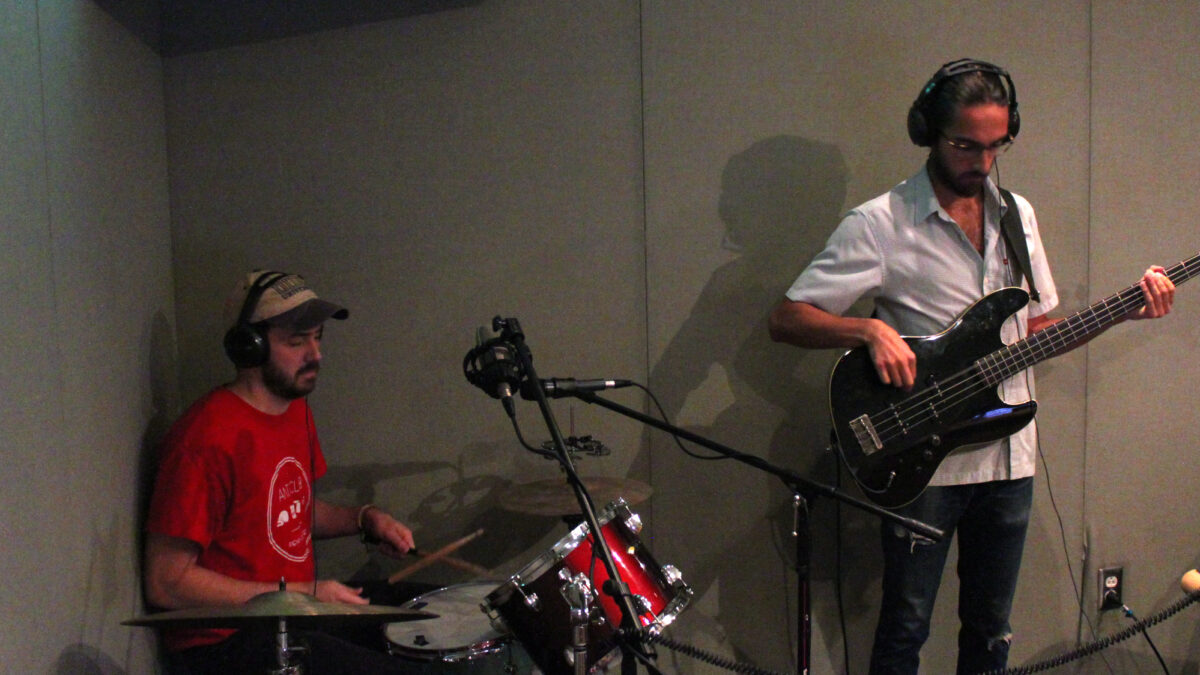 An adult playing drum set and an adult playing electric bass, both wearing headphones in a radio studio