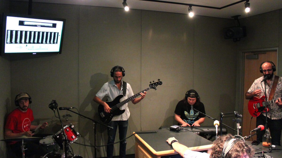 An adult playing drum set, an adult playing electric bass, an adult playing piano keyboard, and an adult playing electric guitar and singing into a microphone, all wearing headphones in a radio studio as another adult wearing headphones listens