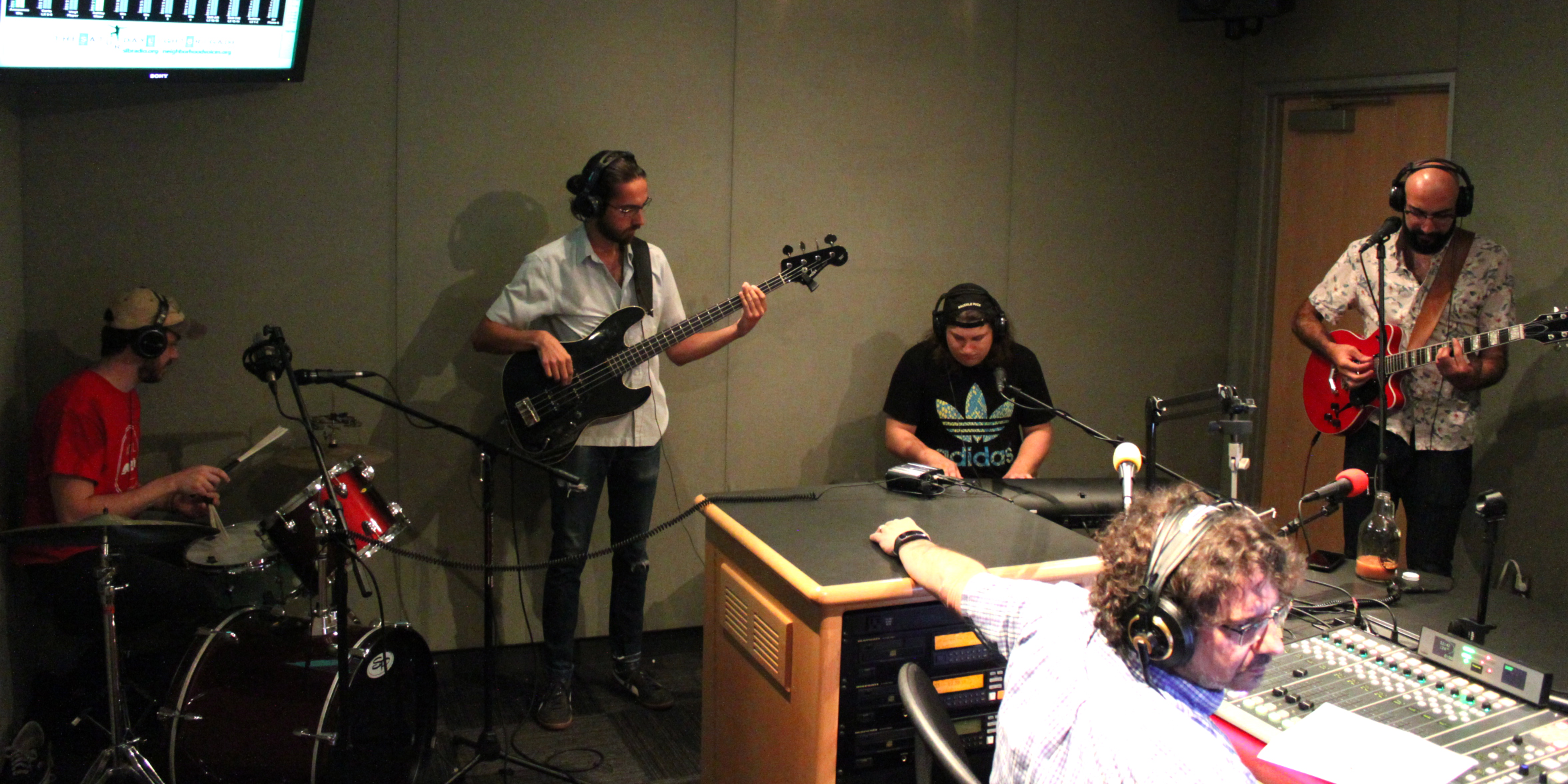 An adult playing drum set, an adult playing electric bass, an adult playing piano keyboard, and an adult playing electric guitar and singing into a microphone, all wearing headphones in a radio studio while an adult wearing headphones listens