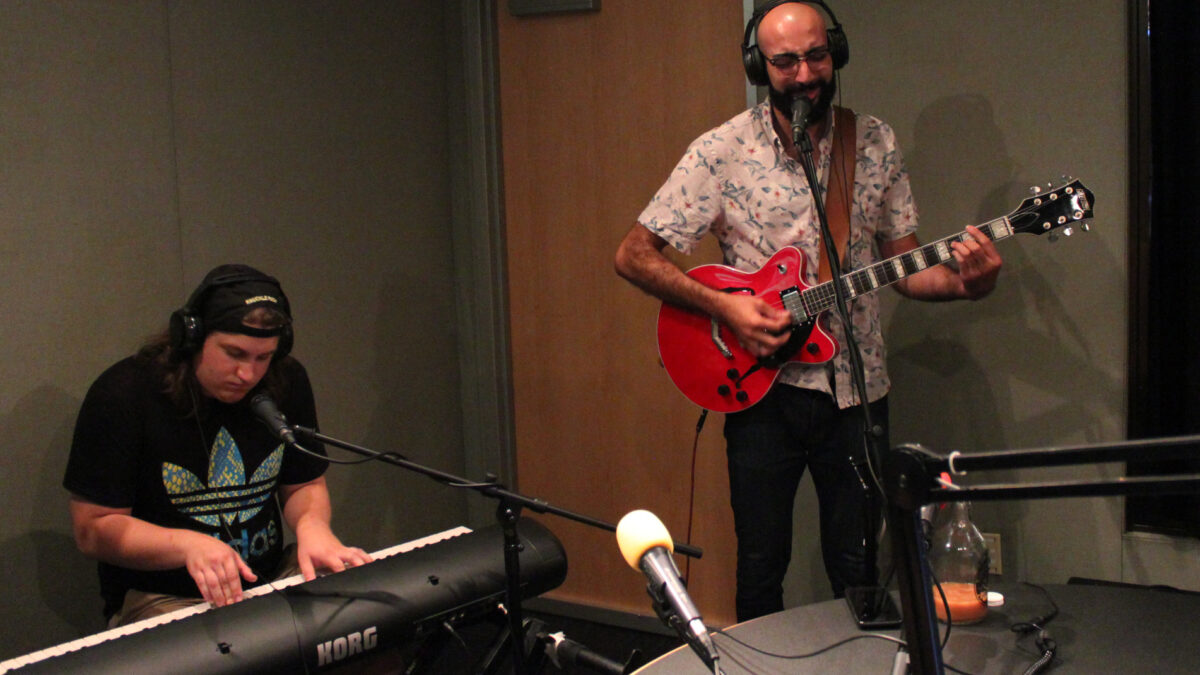 An adult playing piano keyboard wearing headphones and an adult wearing headphones playing electric guitar and singing into a microphone in a radio studio