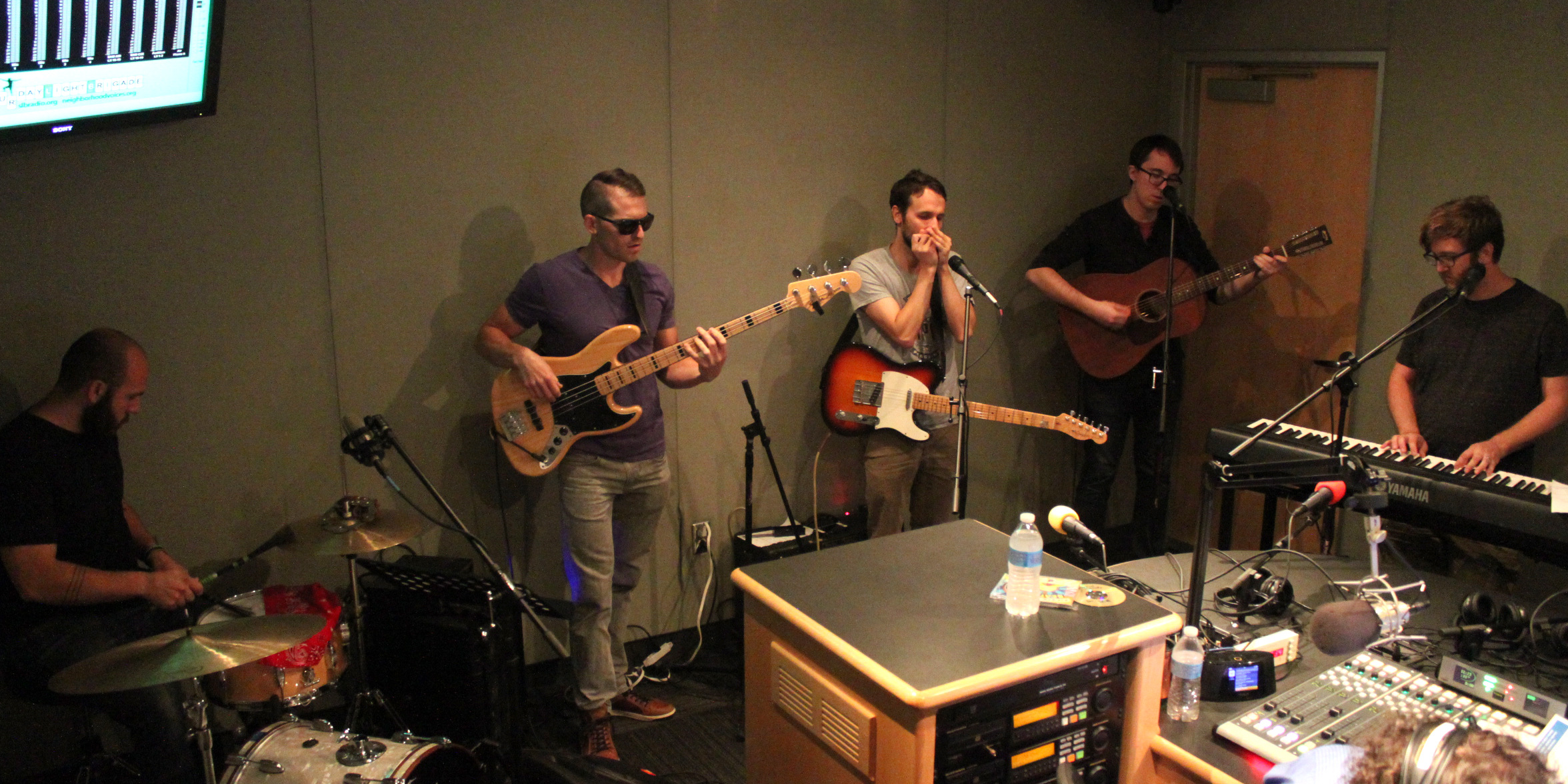 An adult playing drum set, an adult playing bass, an adult holding an electric guitar and playing harmonica into a microphone, an adult playing acoustic guitar and singing into a microphone, and an adult playing piano keyboard in a radio studio.