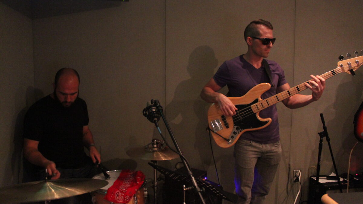 An adult playing drum set and an adult playing bass in a radio studio