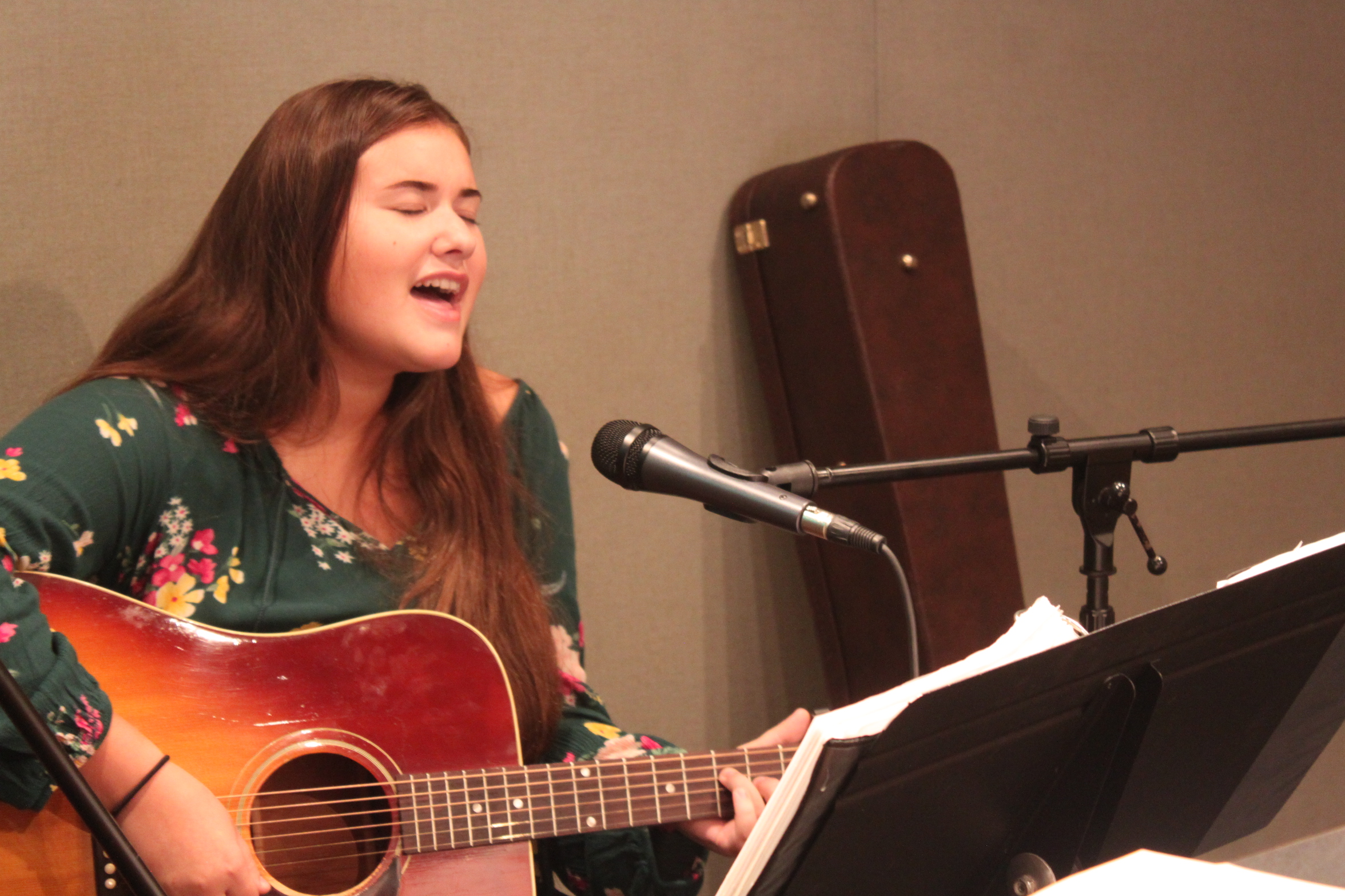 Teen playing guitar and singing into a microphone looking at sheet music on a music stand in a radio studio