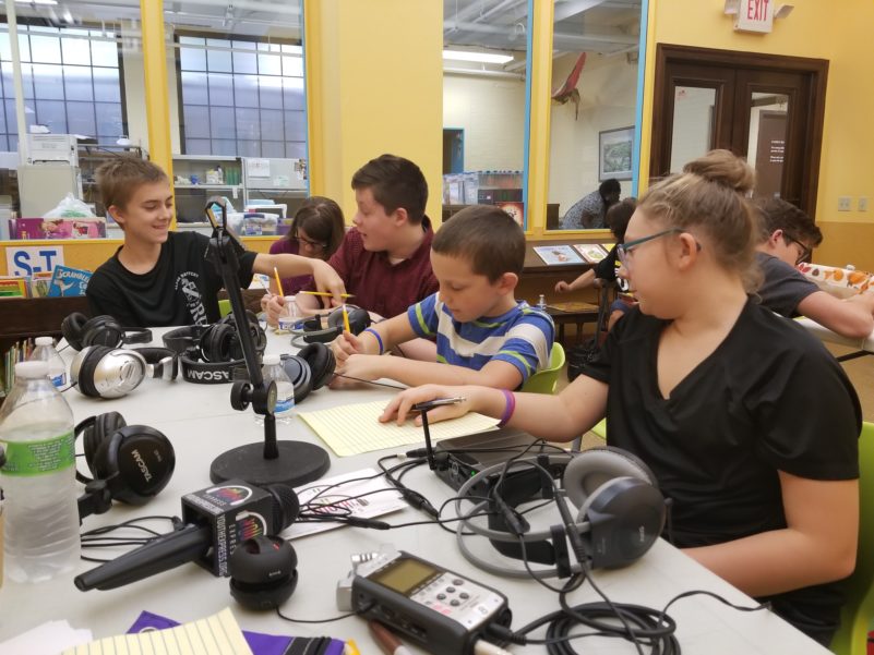 Five youth sitting at a white table covered with headphones, wires, microphones, and a portable recorder