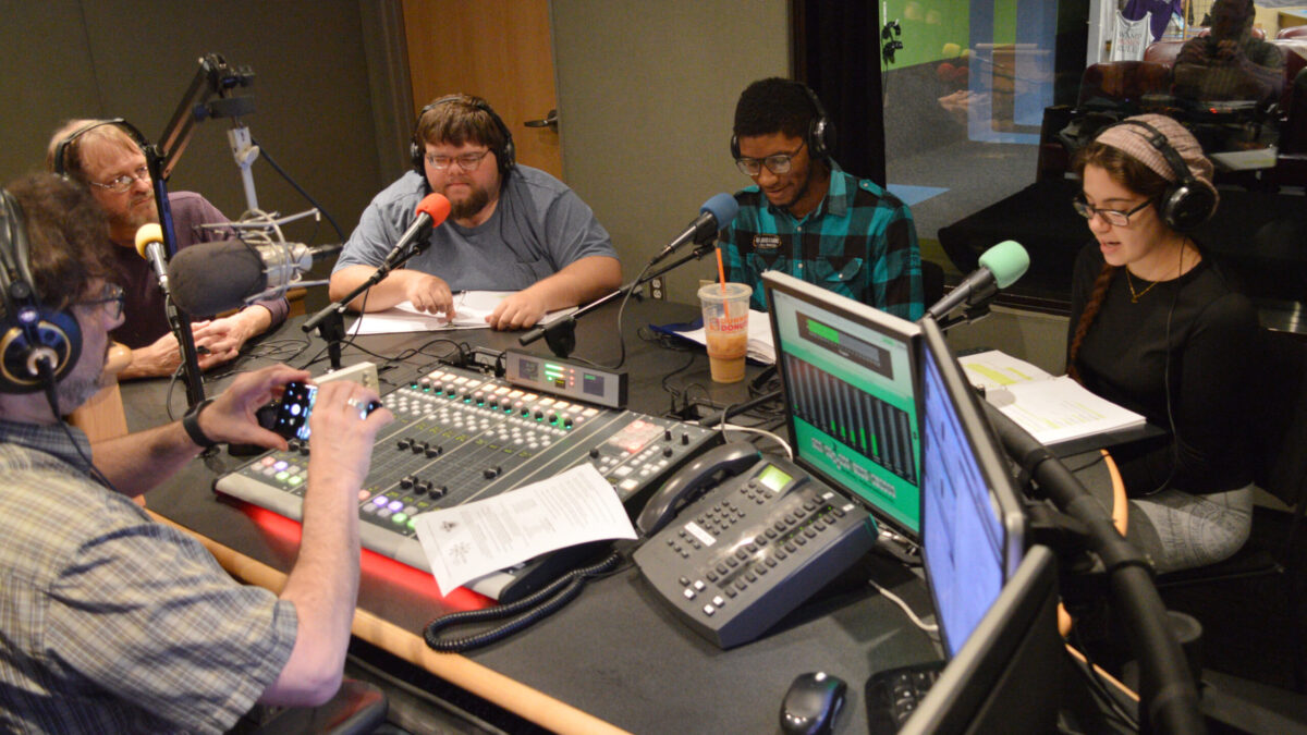 Four adults wearing headphones looking at paper in binders and speaking into microphones in a radio studio while another adult wearing headphones looks at their smartphone