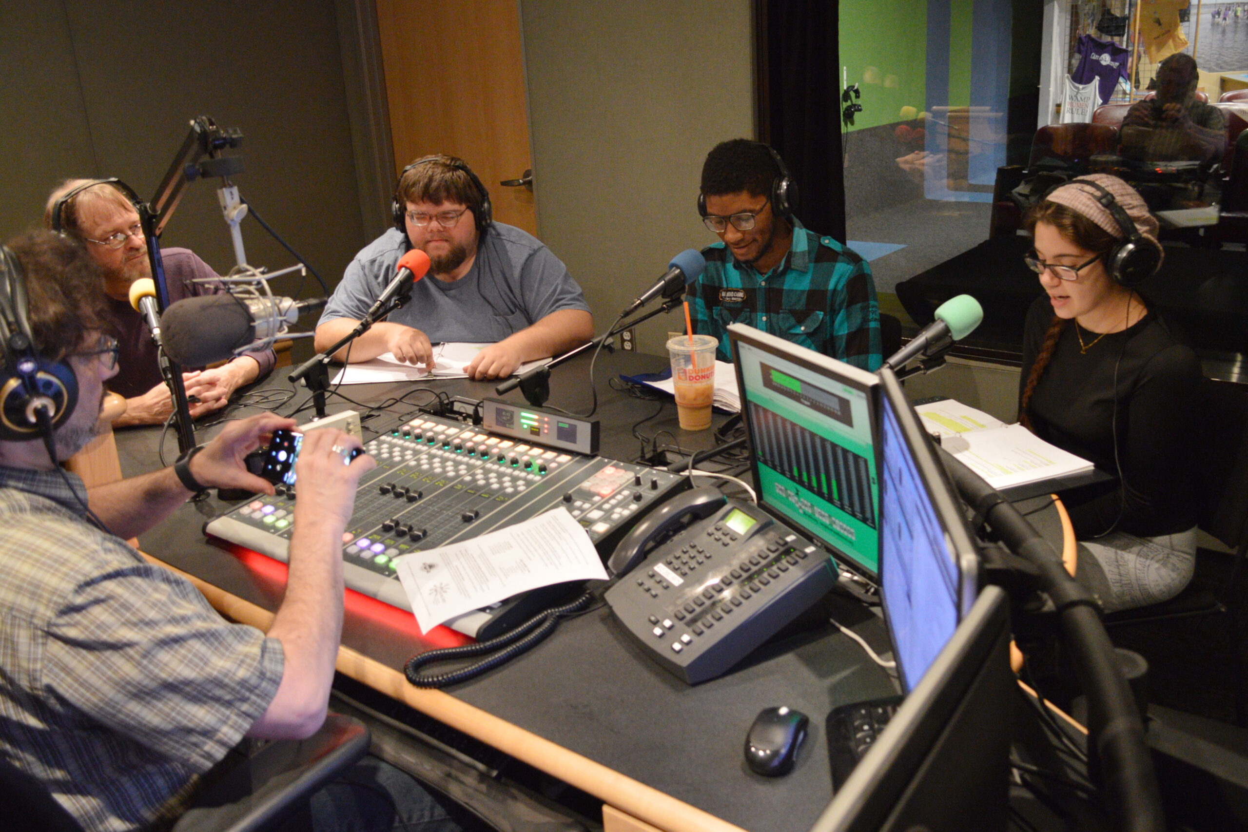 Four adults wearing headphones looking at paper in binders and speaking into microphones in a radio studio while another adult wearing headphones looks at their smartphone