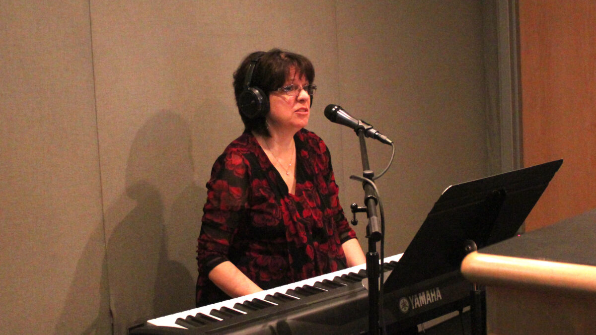 An adult wearing headphones singing into a microphone sitting behind a piano keyboard and a music stand in a radio studio