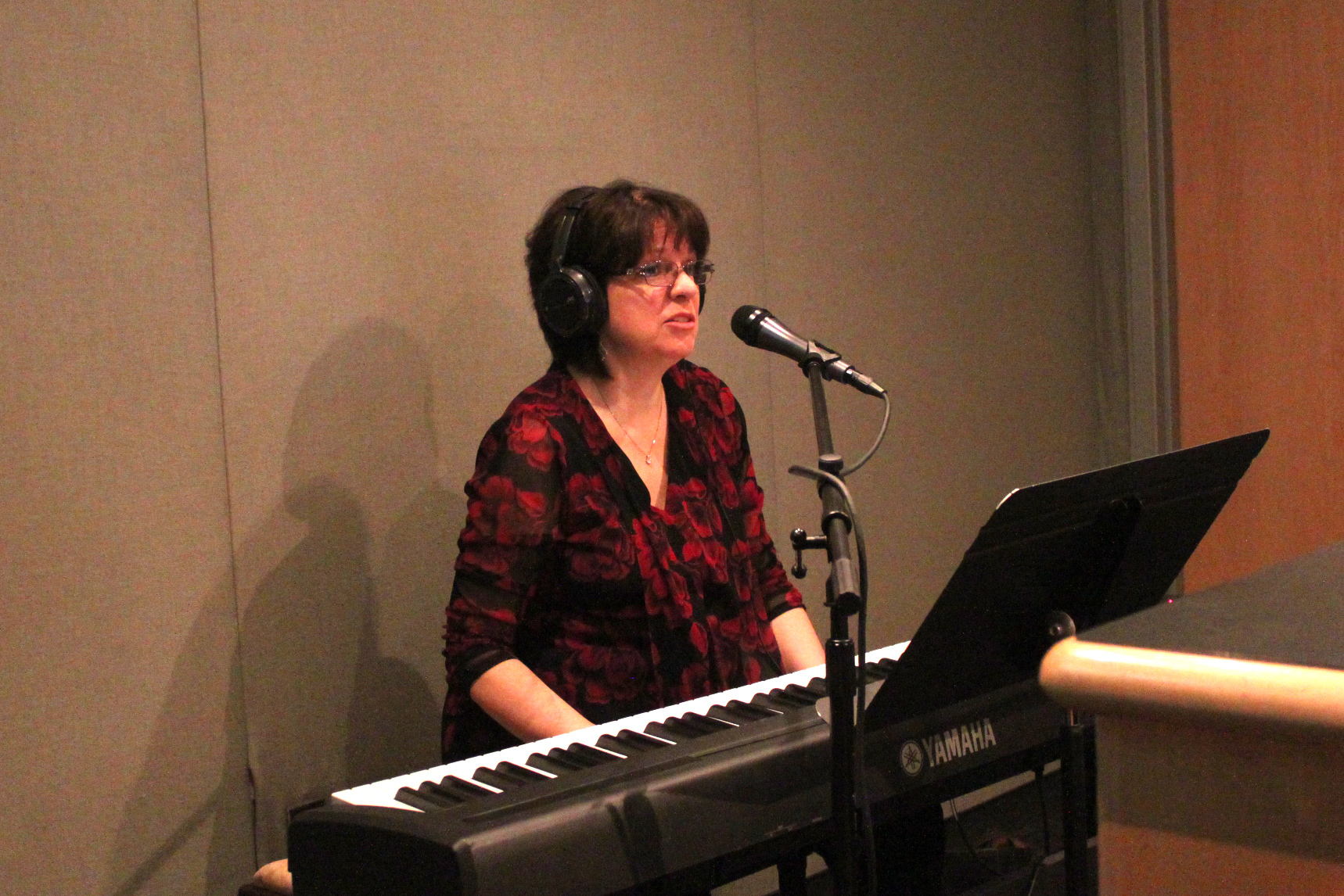 An adult wearing headphones singing into a microphone sitting behind a piano keyboard and a music stand in a radio studio