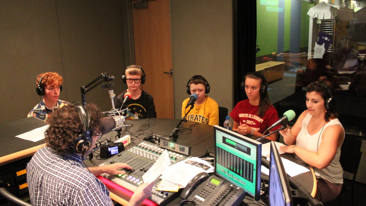 Four youth wearing headphones and an adult wearing headphones sitting behind microphones being interviewed by an adult wearing headphones interacting with a mixing board and holding a piece of paper in a radio studio