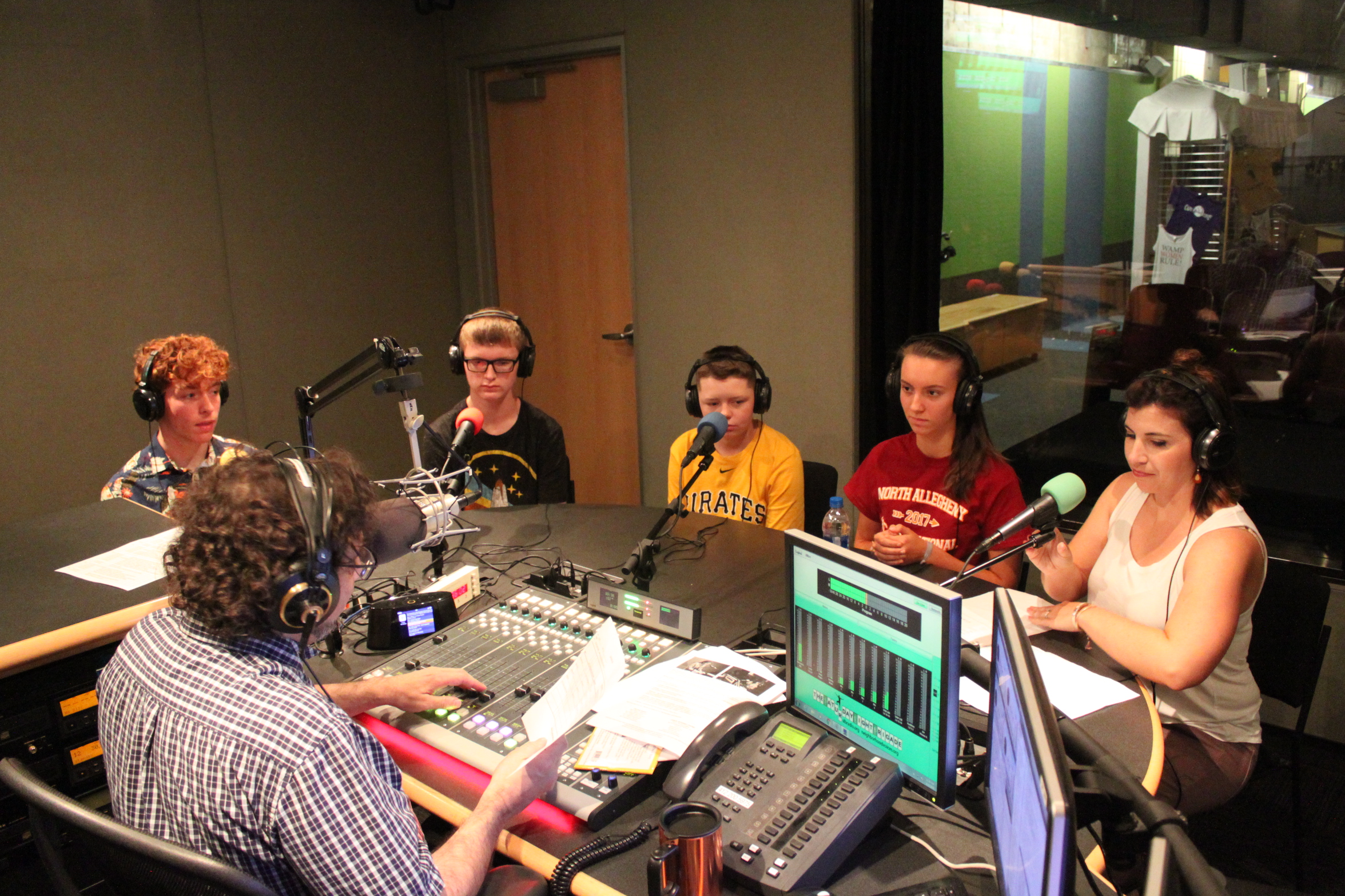 Four youth wearing headphones and an adult wearing headphones sitting behind microphones being interviewed by an adult wearing headphones interacting with a mixing board and holding a piece of paper in a radio studio
