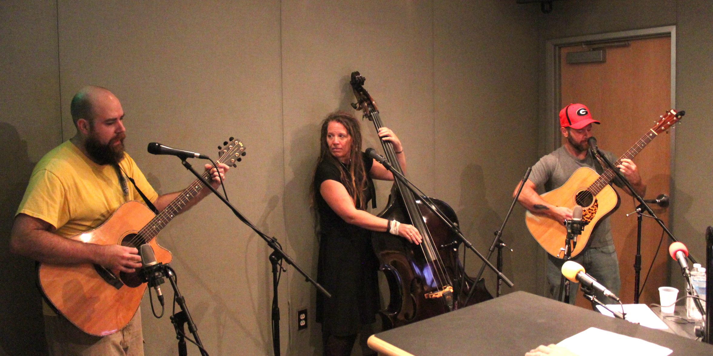 An adult playing guitar and singing into a microphone, an adult playing upright bass, and an adult playing guitar and singing into a microphone in a radio studio