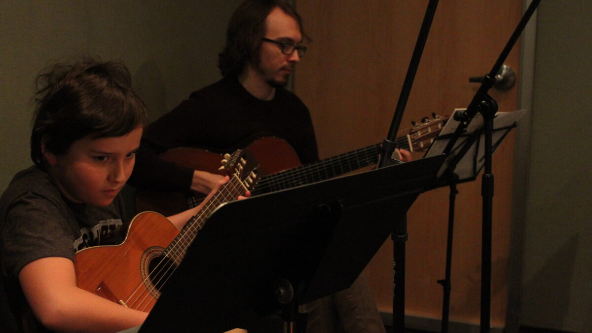 A youth playing guitar and looking at sheet music on a music stand, and an adult playing guitar and looking at sheet music on a music stand in a radio studio