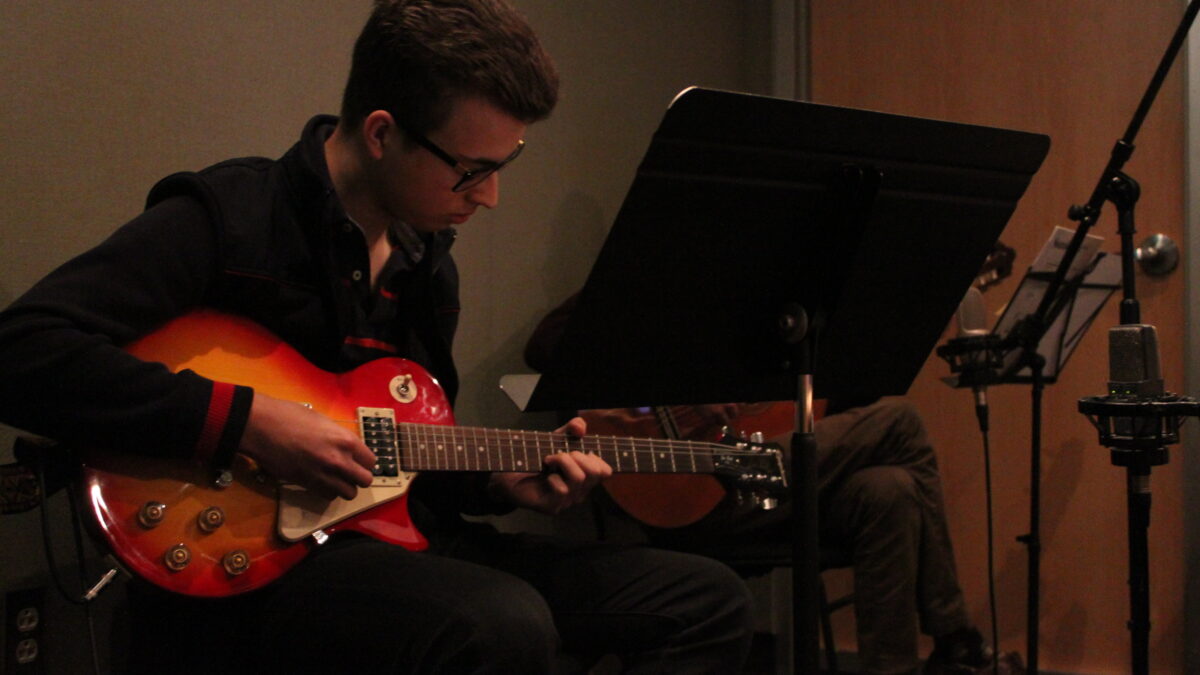 A young adult playing electric guitar and an adult playing acoustic guitar in a radio studio