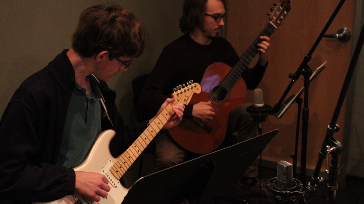 A young adult playing electric guitar and an adult playing acoustic guitar in a radio studio