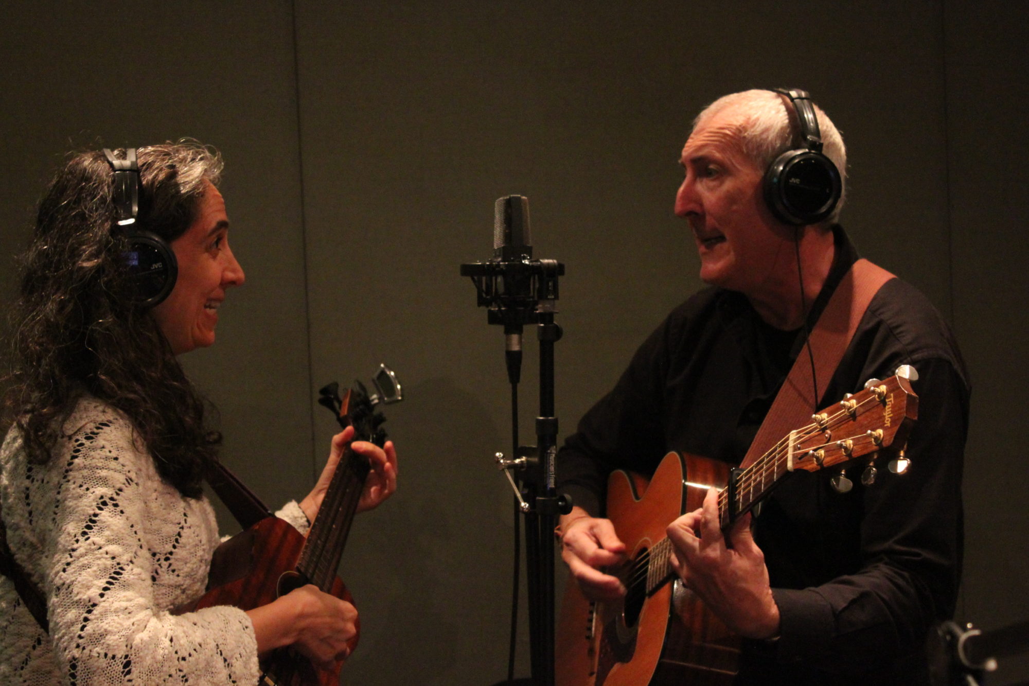Two adults wearing headphones singing into a microphone in a radio studio, one playing ukulele, the other playing guitar