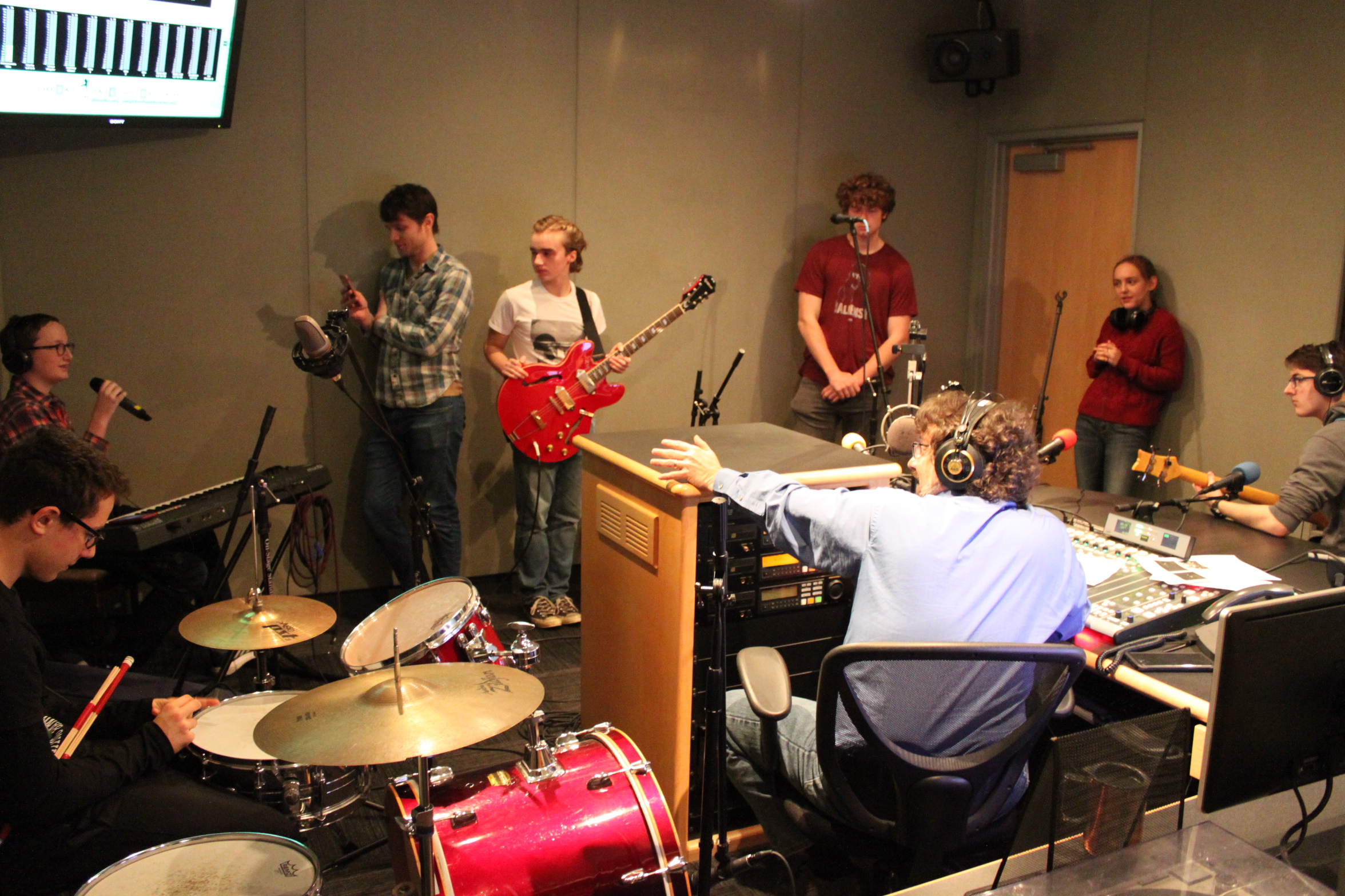 A teen sitting at a drum set, a teen sitting behind a piano keyboard speaking into a microphone, an individual holding a smartphone, a teen holding a guitar, teen standin in a corner, a teen leaning against a wall, and a teen wearing headphones sitting while holding a bass, all being interviewed by an adult wearing headphones sitting behind a mixing board in a radio studio