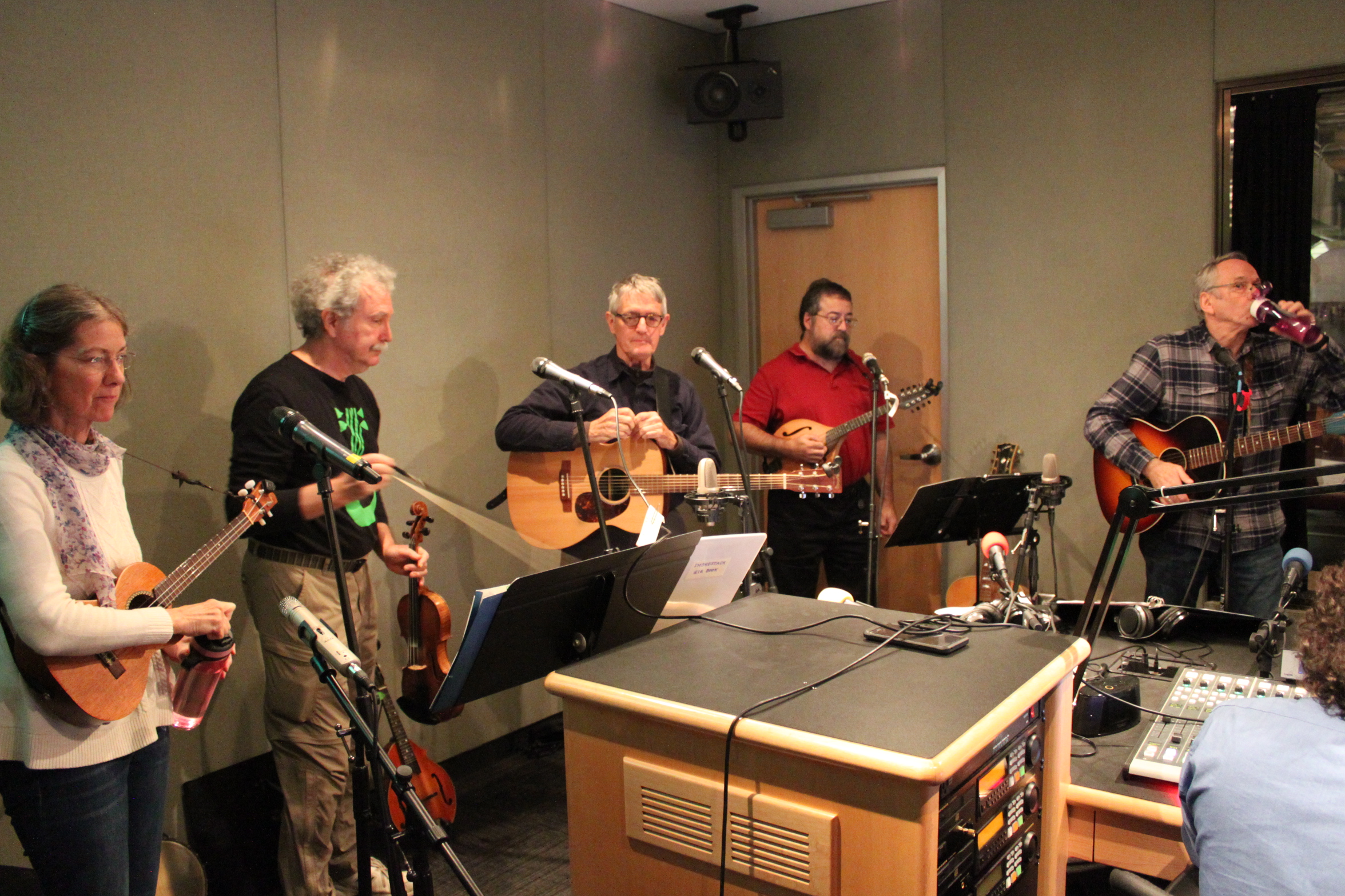 An adult holding a ukulele and a water bottle, an adult holding a violin, an adult holding a guitar, an adult holding a mandolin, and an adult holding a guitar drinking from a plastic water bottle in a radio studio
