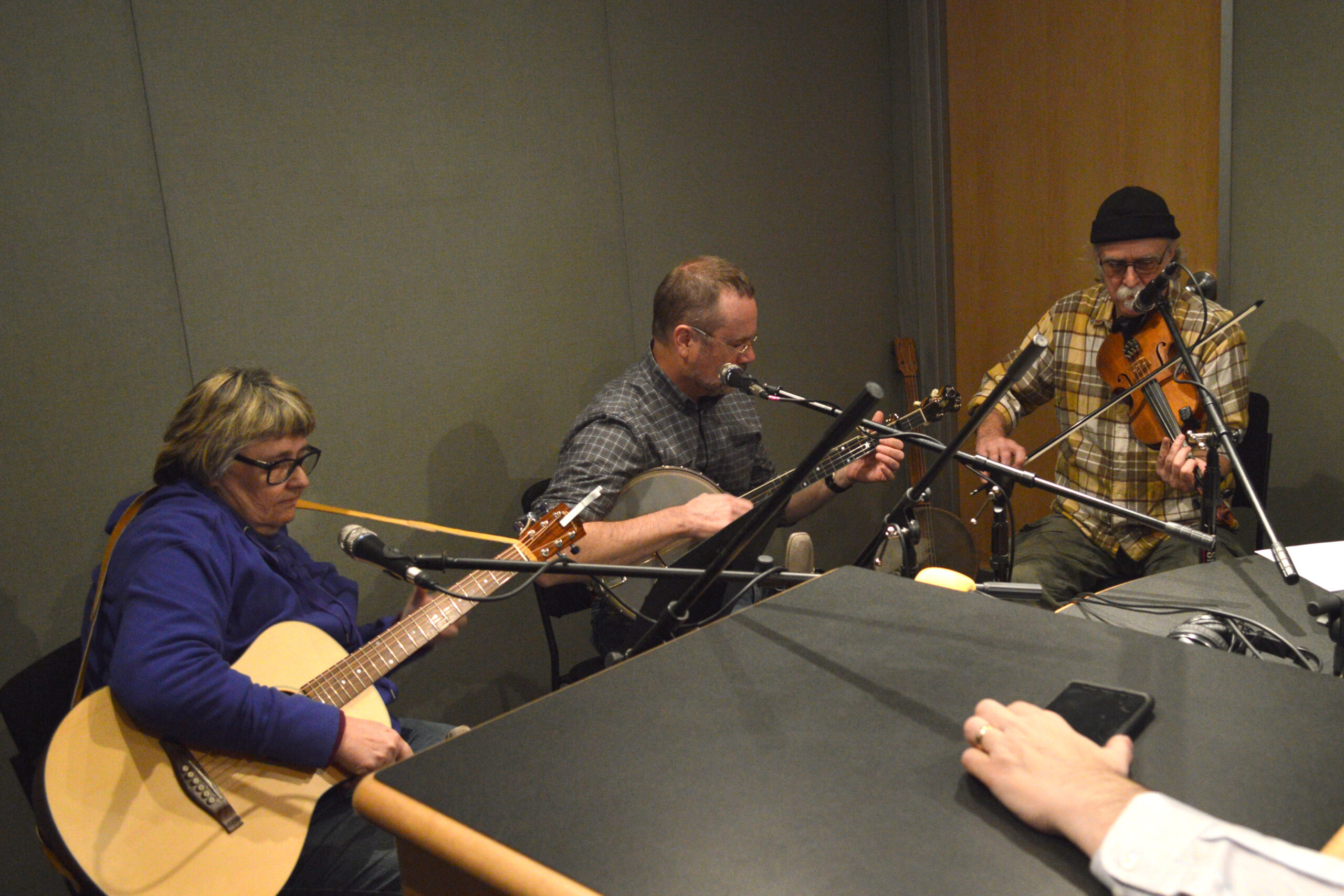An adult playing guitar, an adult playing banjo, and an adult playing violin, all sitting behind microphones in a radio studio