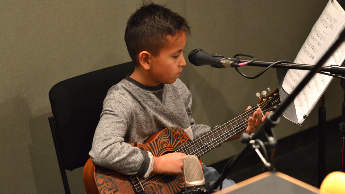 Child playing guitar sitting behind microphones and looking at sheet music on a music stand in a radio studio