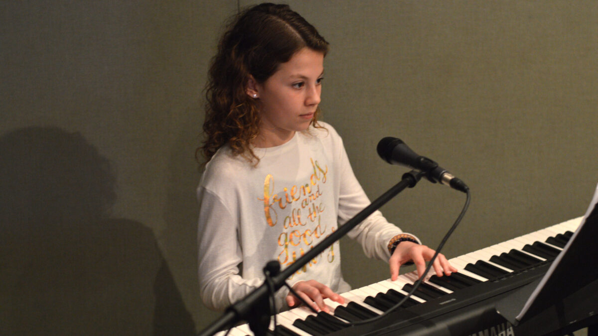 A child sitting behind a microphone playing piano looking at sheet music on a music stand in a radio studio
