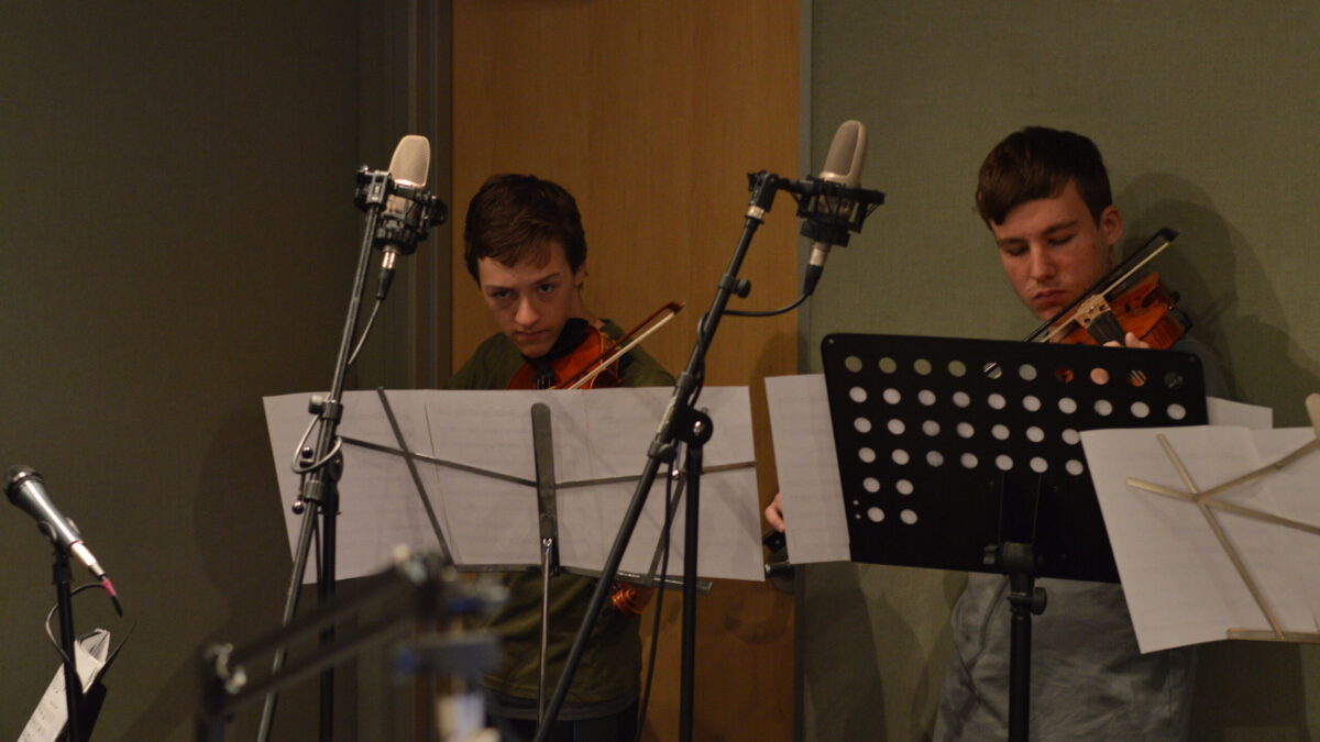 Two teens playing violin looking at sheet music on music stands in a radio studio
