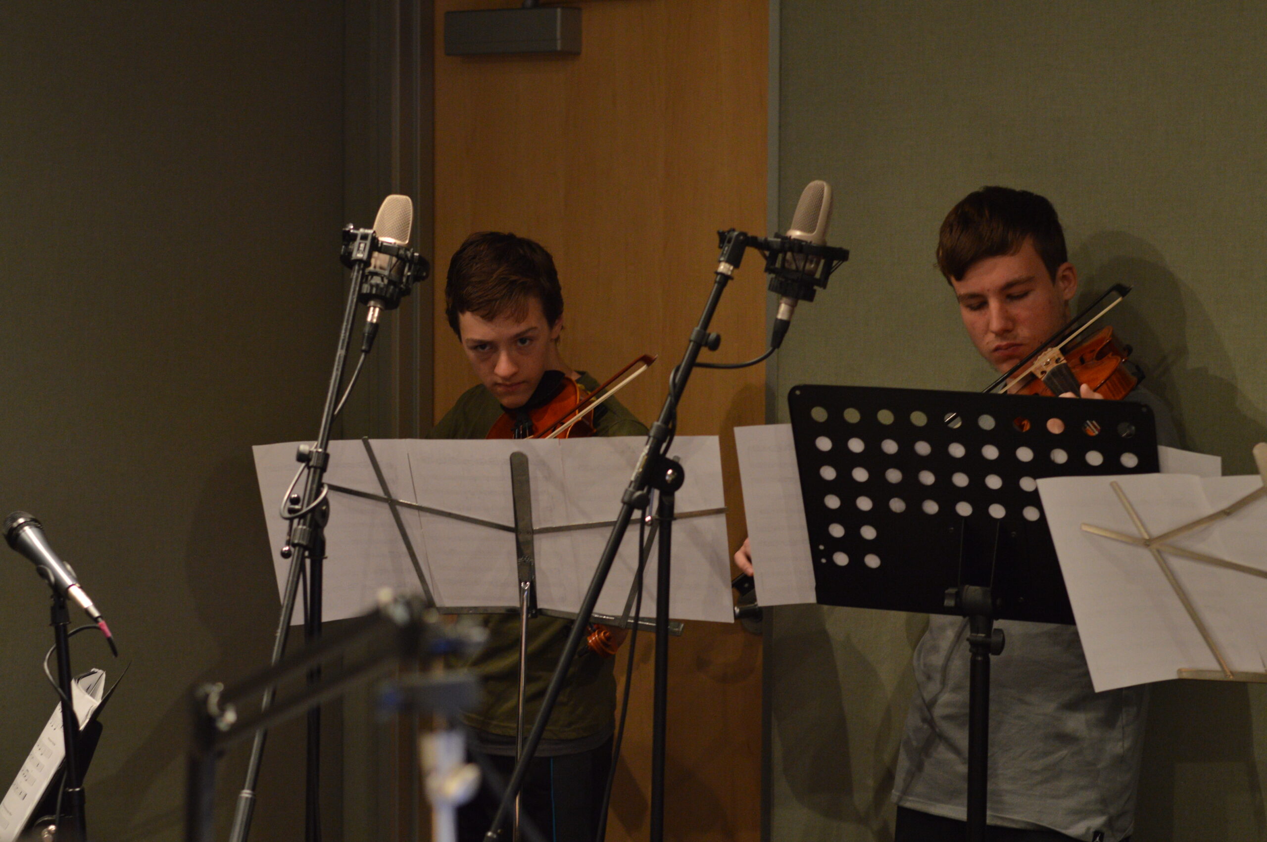 Two teens playing violin looking at sheet music on music stands in a radio studio