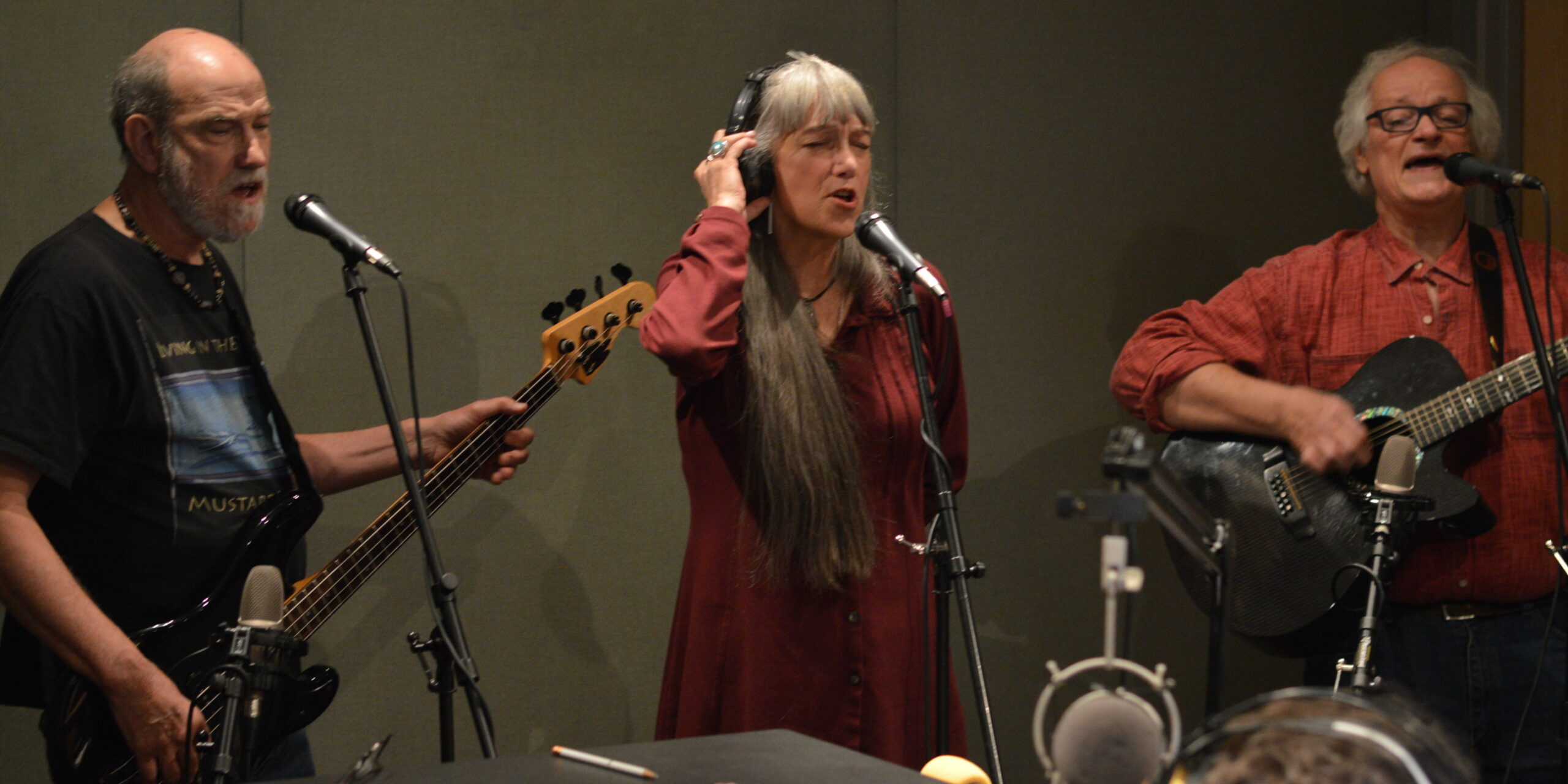 Three adults singing into microphones, one playing bass, another playing guitar, in a radio studio