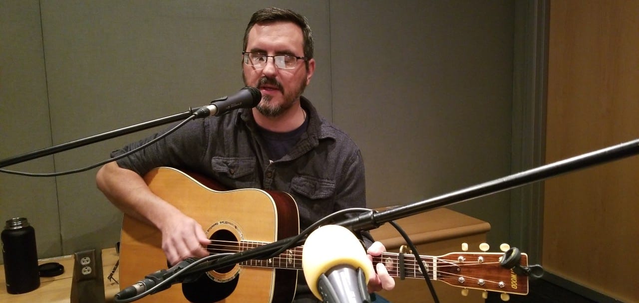 Adult playing guitar and singing into a microphone in a radio studio