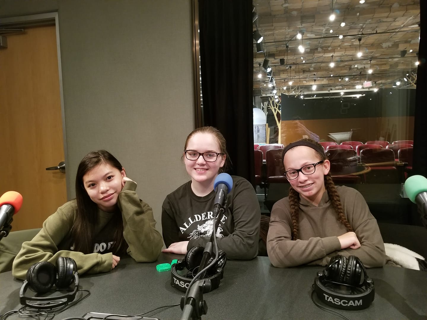 Three teens smiling sitting behind microphones and headphones in a radio studio