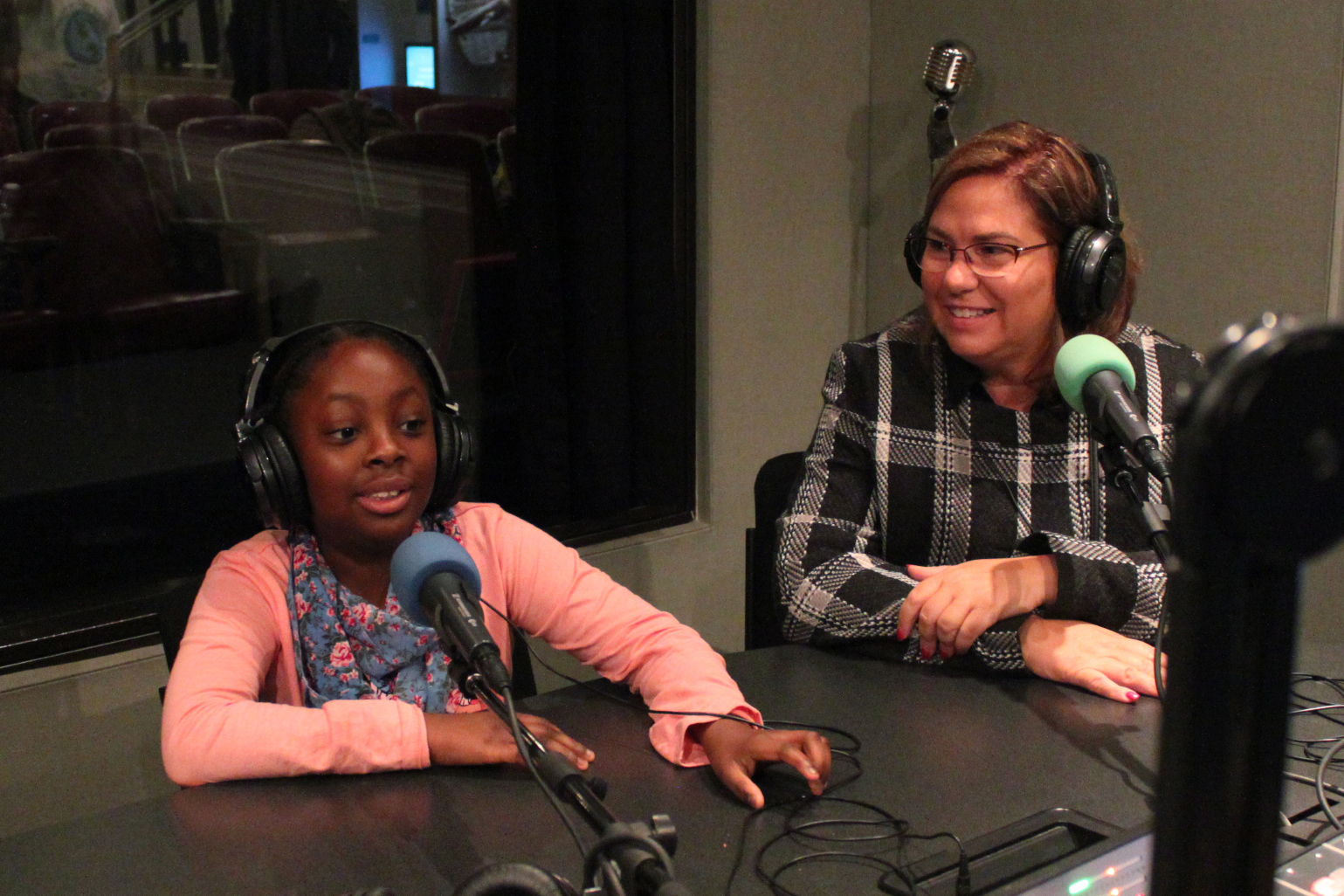 A child wearing headphones speaking into a microphone and an adult wearing headphones sitting behind a microphone in a radio studio