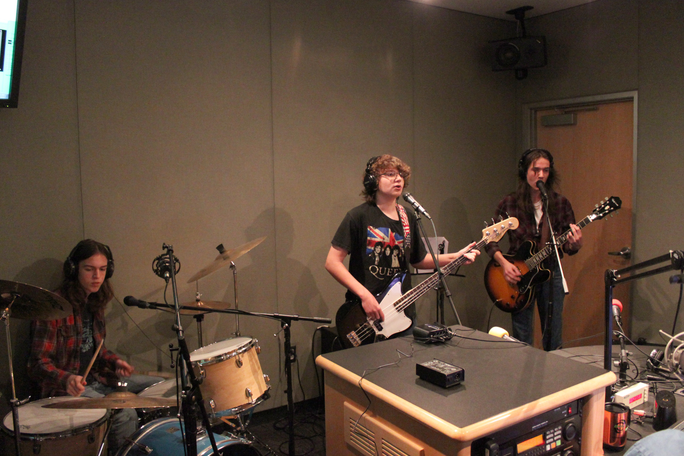 A teen wearing headphones playing a drum set, a teen wearing headphones playing bass and singing into a microphone, and a teen wearing headphones playing guitar and singing into a microphone in a radio studio