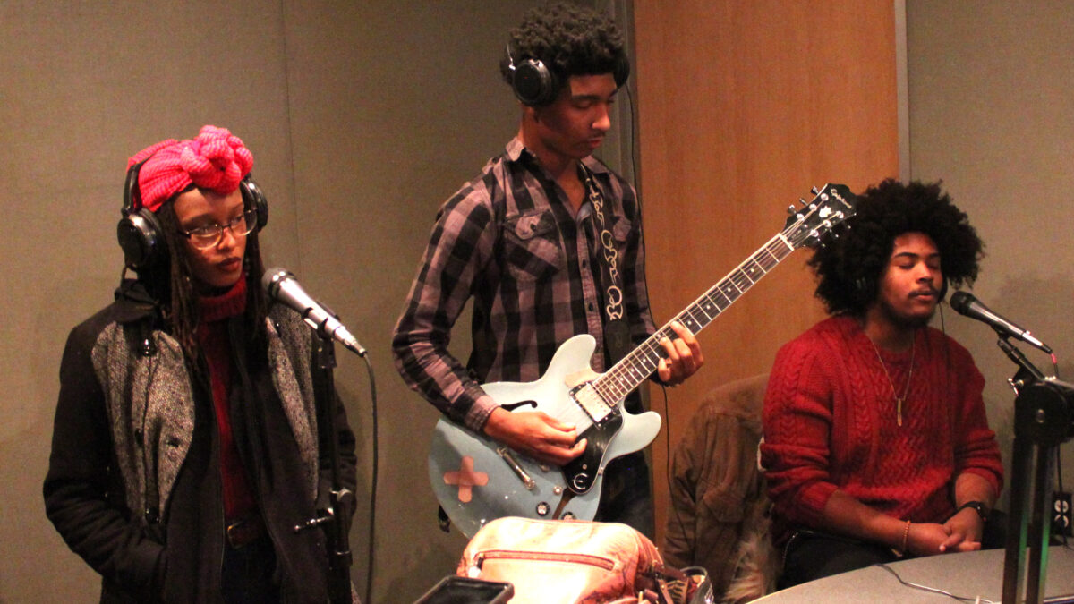 Three teens wearing headphones, two singing into microphones, one playing guitar, in a radio studio