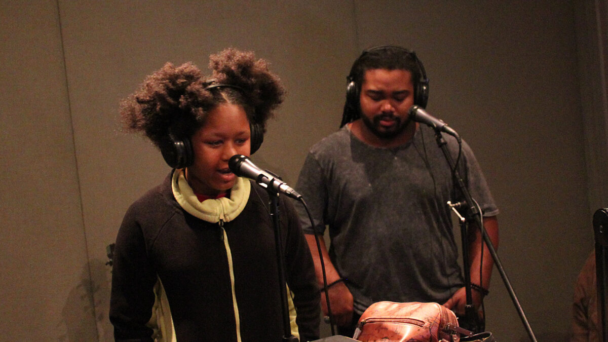 Youth wearing headphones singing into a microphone and an adult wearing headphones standing behind a microphone in a radio studio