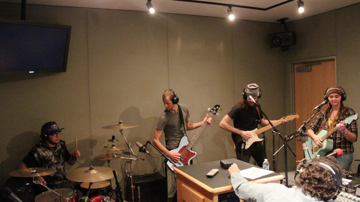Four young adults wearing headphones performing on drumset, bass, electric guitar, and acoustic guitar for another adult wearing headphones sitting behind a mixing board in a radio studio