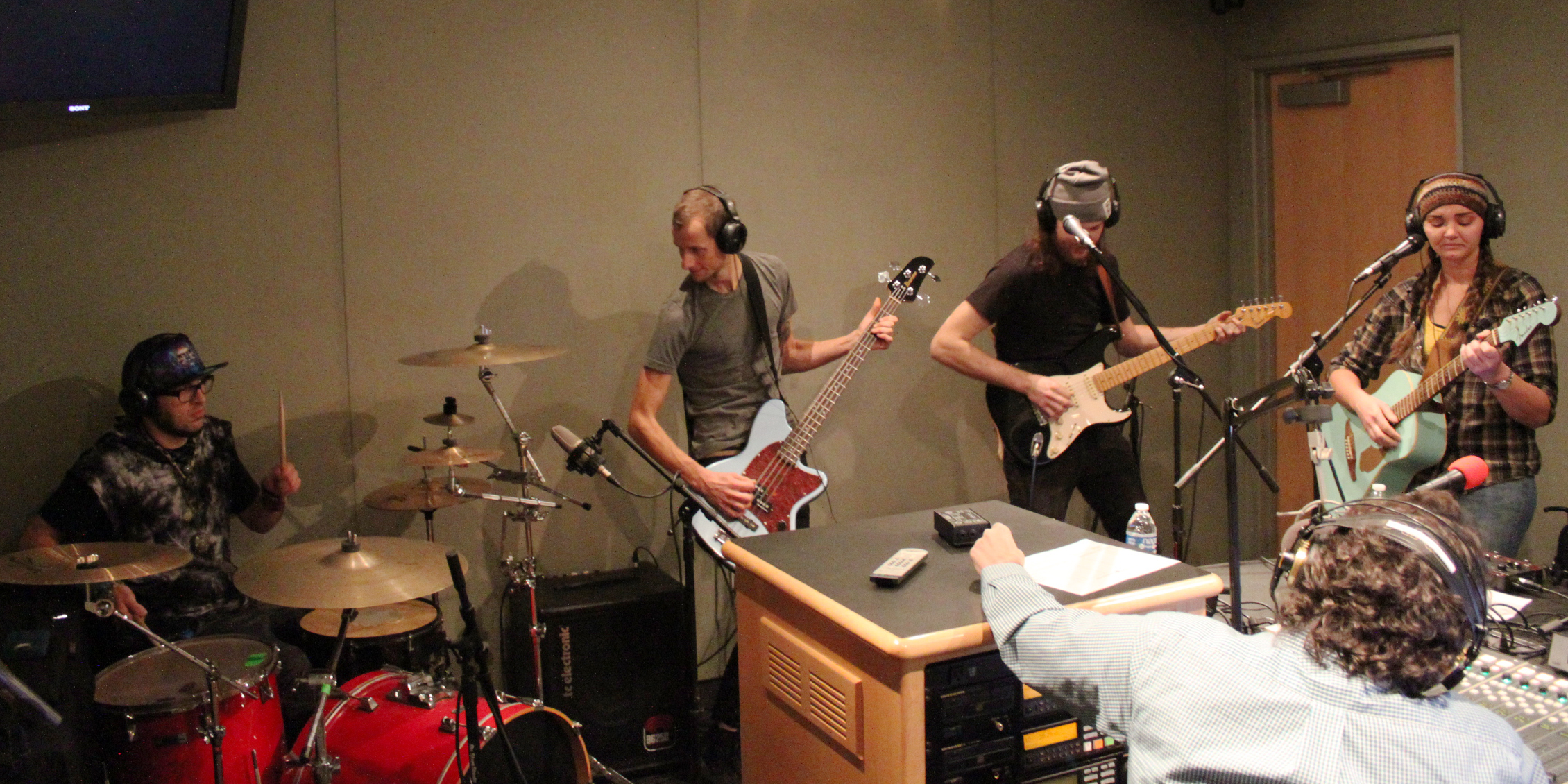 Four young adults wearing headphones performing on drumset, bass, electric guitar, and acoustic guitar for another adult wearing headphones sitting behind a mixing board in a radio studio