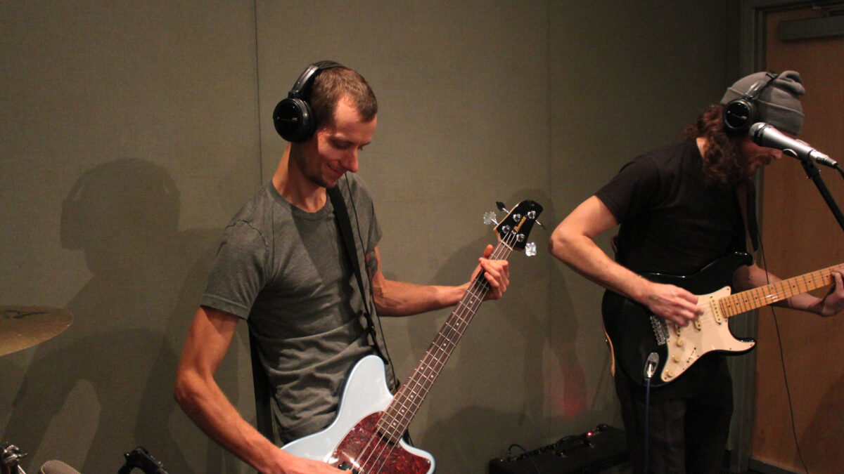 Two young adults wearing headphones in a radio studio, one playing bass, the other playing electric guitar