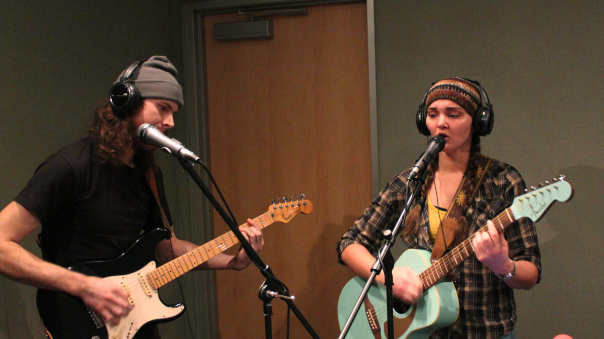 Two young adults wearing headphones and singing into microphone in a radio studio, one playing electric guitar, the other playing acoustic guitar