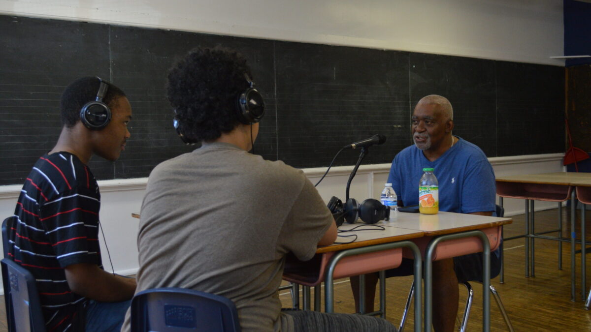Two teens wearing headphones interviewing an adult speaking into a microphone on two classroom desks in a classroom