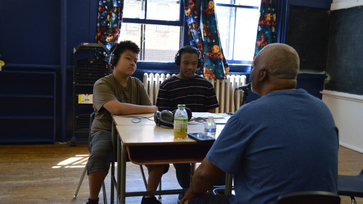 Two teens wearing headphones interviewing an adult speaking into a microphone connected to a portable recorder on two classroom desks in a classroom