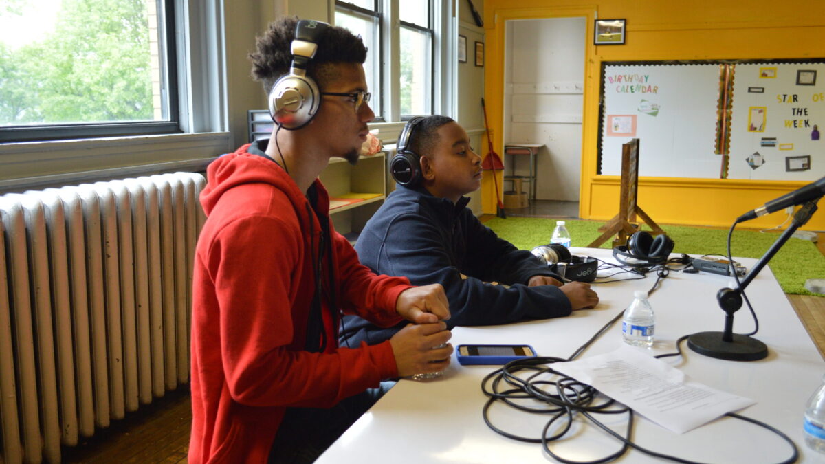 Two teens wearing headphones looking at a microphone connected to a portable recorder on a white table in a classroom