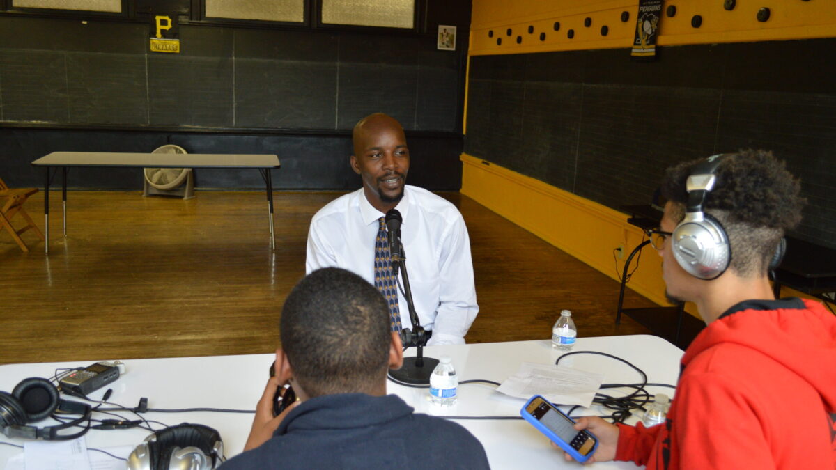 Two teens wearing headphones interviewing an adult speaking into a microphone connected to a portable recorder on a white table in a classroom